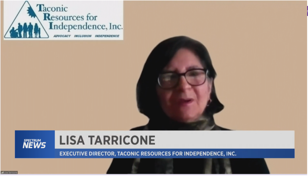 Video image of Lisa Tarricone, Executive Director of Taconic Resources for Independence, Inc. (TRI) interviewed by Spectrum News 1.  Behind Lisa is the TRI logo in the upper left corner. Lisa is a white female, black hair, wearing glasses, a black sweater, and scarf.